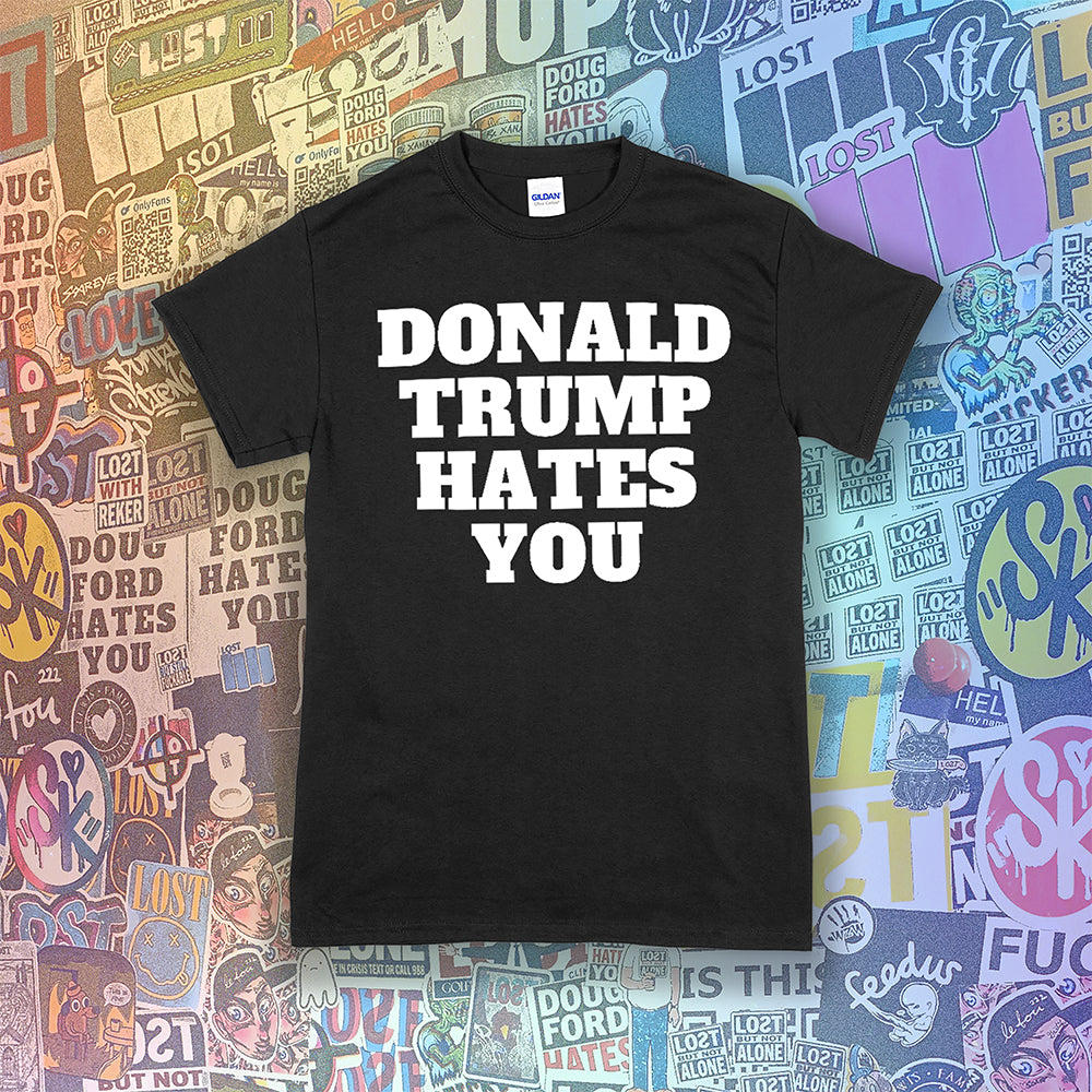 The Donald Trump Hates You t-shirt. Bold letters screen printed with white ink on a black shirt. Colorful assorted graffiti stickers are in the background.