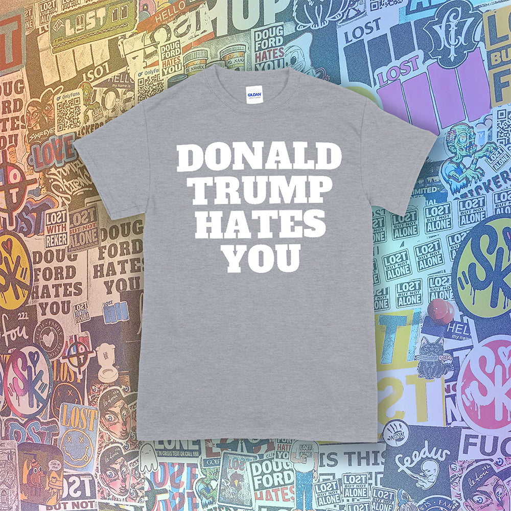 The Donald Trump Hates You t-shirt. Bold letters screen printed with white ink on a grey shirt. Colorful assorted graffiti stickers are in the background.