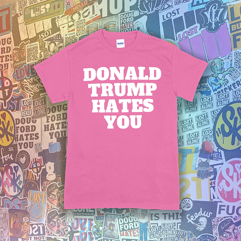 The Donald Trump Hates You t-shirt. Bold letters screen printed with white ink on a pink shirt. Colorful assorted graffiti stickers are in the background.