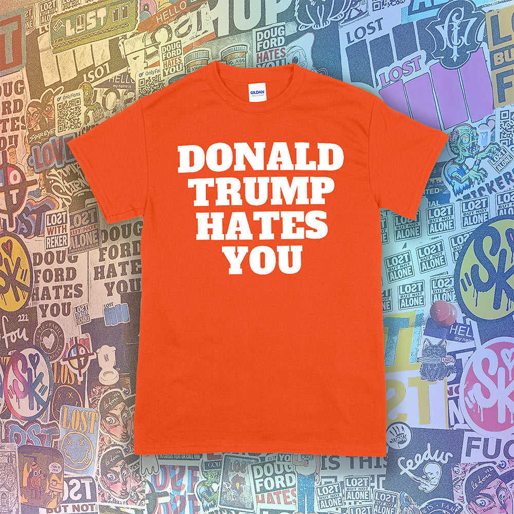 The Donald Trump Hates You t-shirt. Bold letters screen printed with white ink on an orange shirt. Colorful assorted graffiti stickers are in the background.