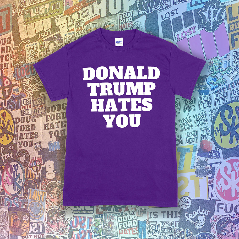 The Donald Trump Hates You t-shirt. Bold letters screen printed with white ink on a purple shirt. Colorful assorted graffiti stickers are in the background.