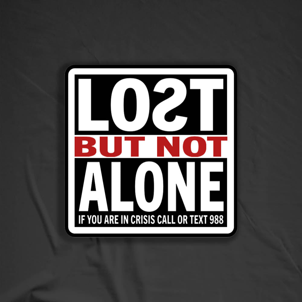 The Lost But Not Alone Sticker by artist and mental health advocate Christopher Reilly. This squared sticker features text stating Lost But Not Alone, if you are in crisis call or text 988. The black, white and red sticker design promotes mental health awareness and suicide prevention. A back background provides contrast for the displayed sticker.