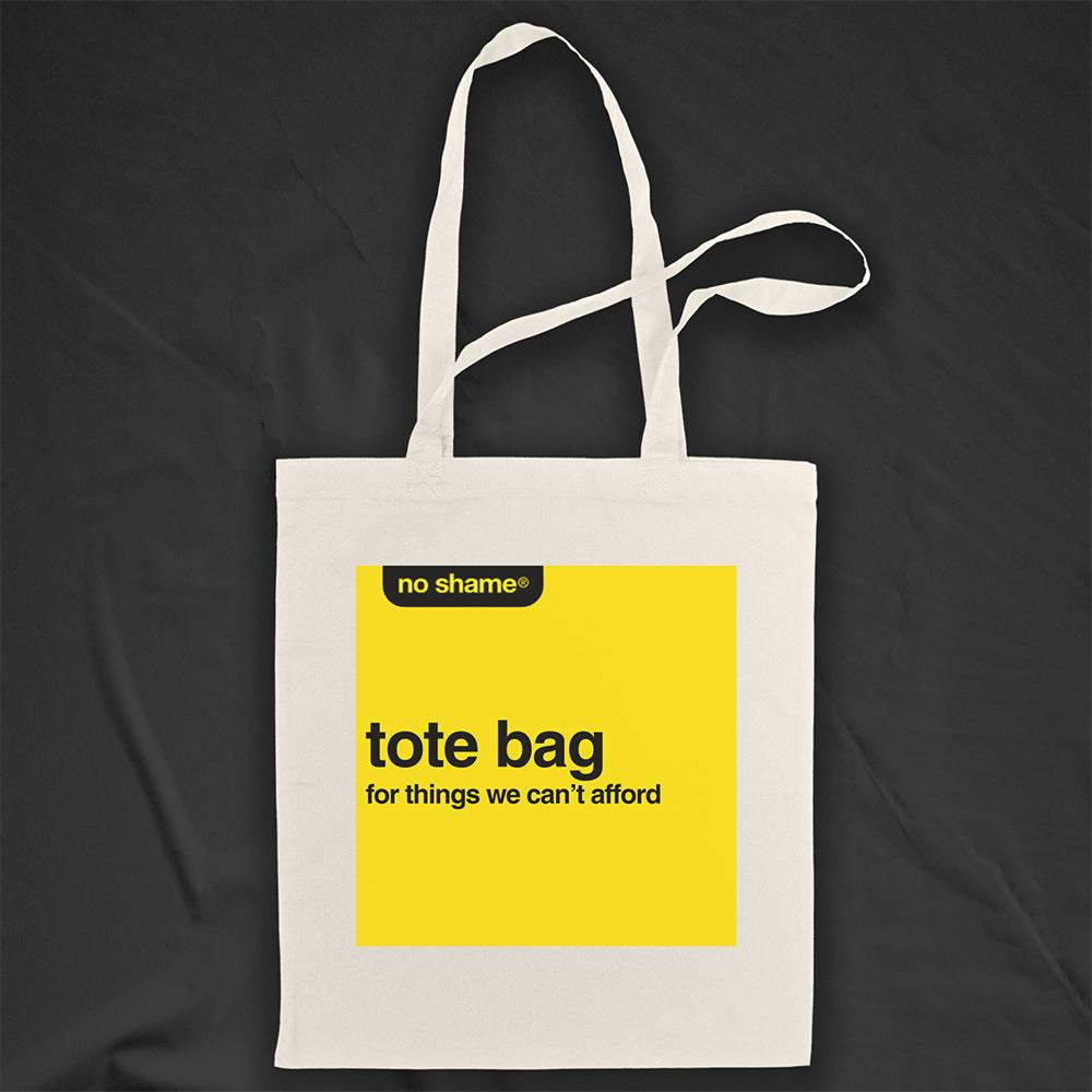 The no shame tote bag: for things we can't afford. Those words are screen  printed in bold black ink inside a no name yellow box on a large, natural canvas tote bag. A black background provides dramatic contrast for the tote bag on display.