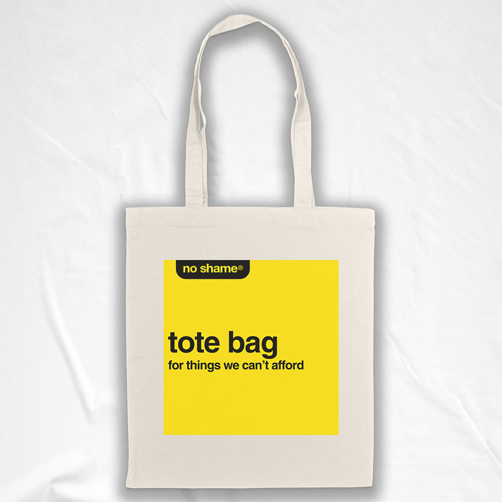The no shame tote bag: for things we can't afford. Those words are screen  printed in bold black ink inside a no name yellow box on a large, natural canvas tote bag. A plain white background provided a distraction-free backdrop for the tote bag on display.