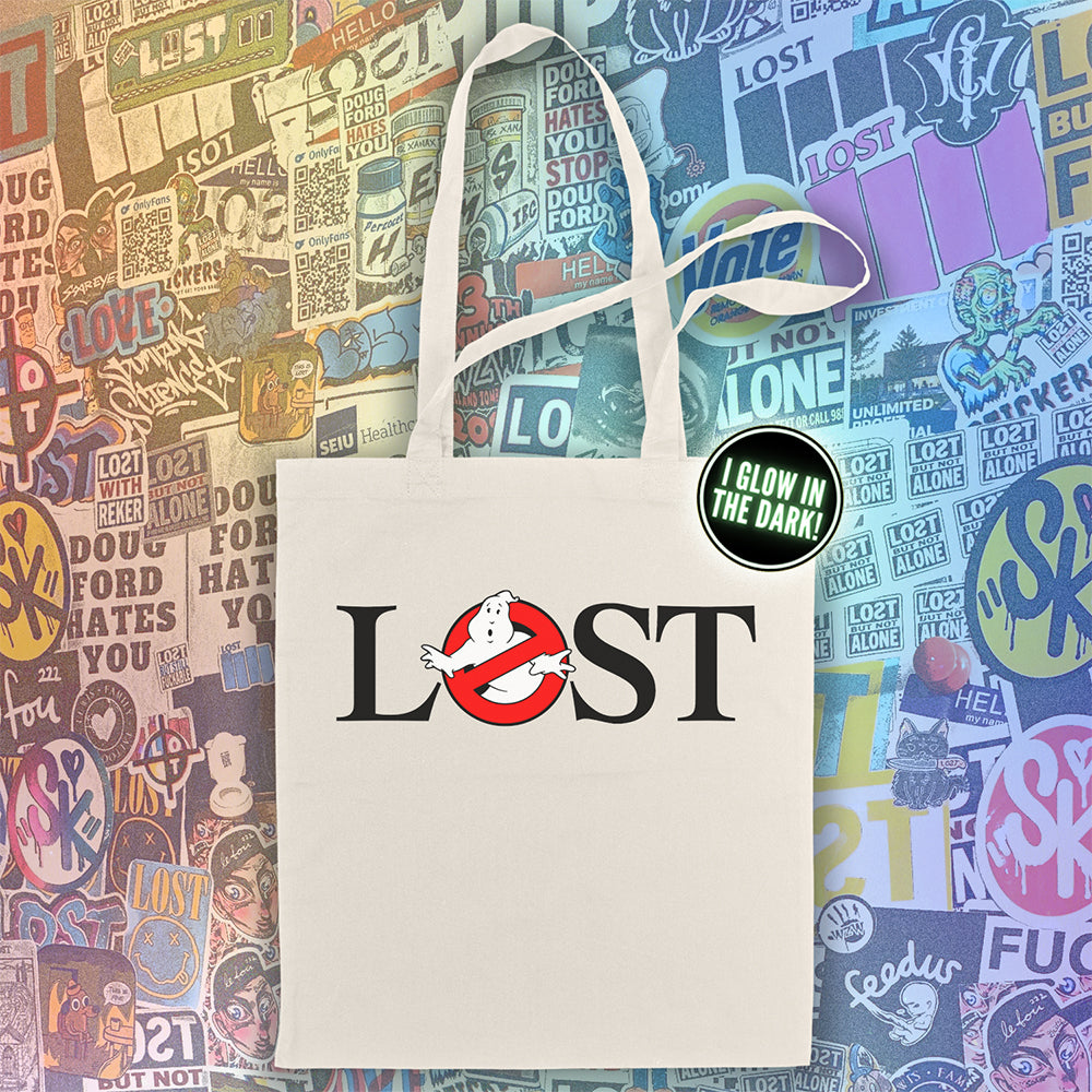 The LOSTBUSTERS tote bag features a surprised looking  ghost trapped in the O of the word LOST and vaguely resembling the original Ghostbusters logo. The crisp image is screen printed on a natural canvas tote bag. A wall of colorful graffiti stickers creates a vibrant background for the tote bag on display.