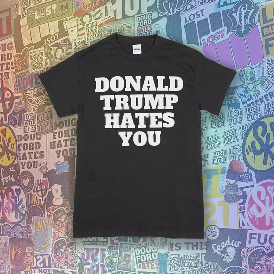 The Donald Trump Hates You t-shirt. Bold letters screen printed with white ink displayed on an assortment of colored shirts. Variety of different graffiti stickers as a background.