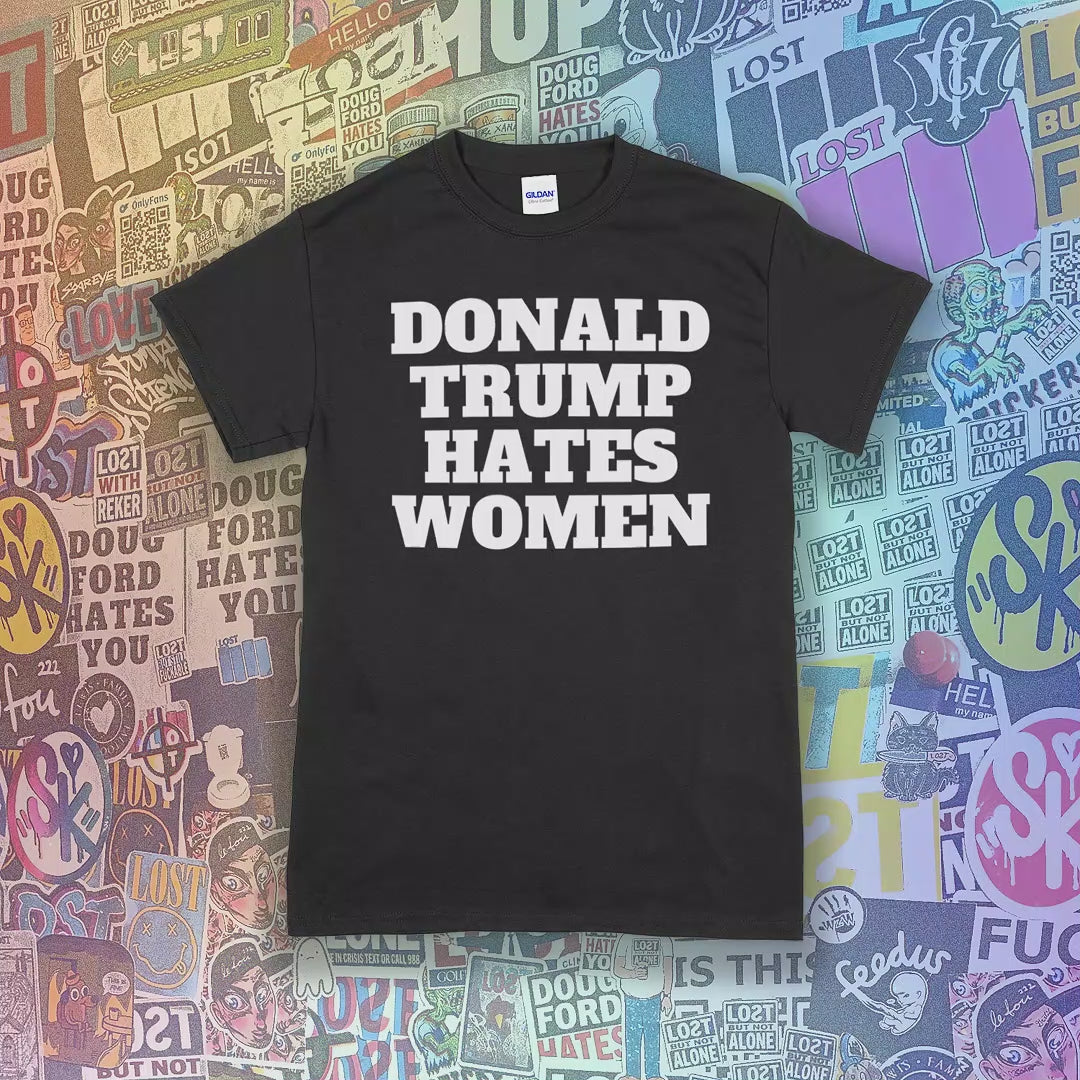 The Donald Trump Hates Women t-shirt. Bold letters screen printed with white ink on a variety of different colored shirts. A colorful variety of graffiti stickers are in the background.